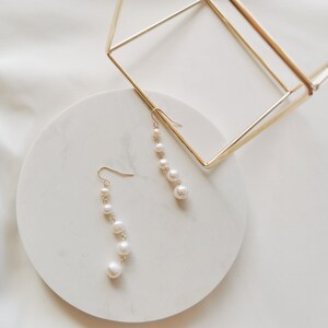 May include: A pair of gold dangle earrings with white pearl beads. The earrings are displayed on a white circular surface.