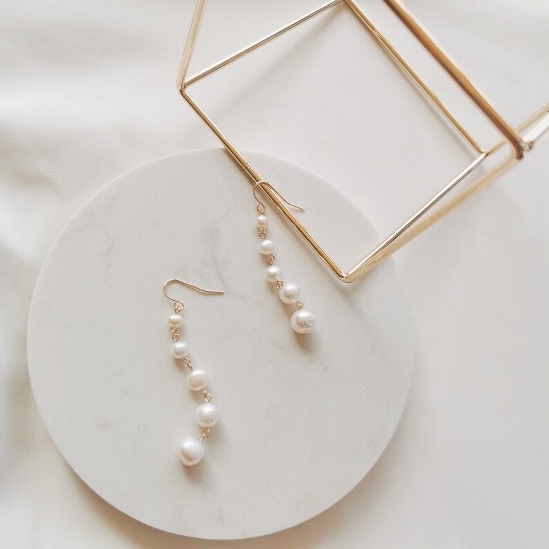 May include: A pair of gold dangle earrings with white pearl beads. The earrings are displayed on a white circular surface.