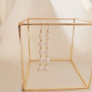 May include: A pair of gold dangle earrings with a chain of white pearls. The earrings are hanging from a gold stand.