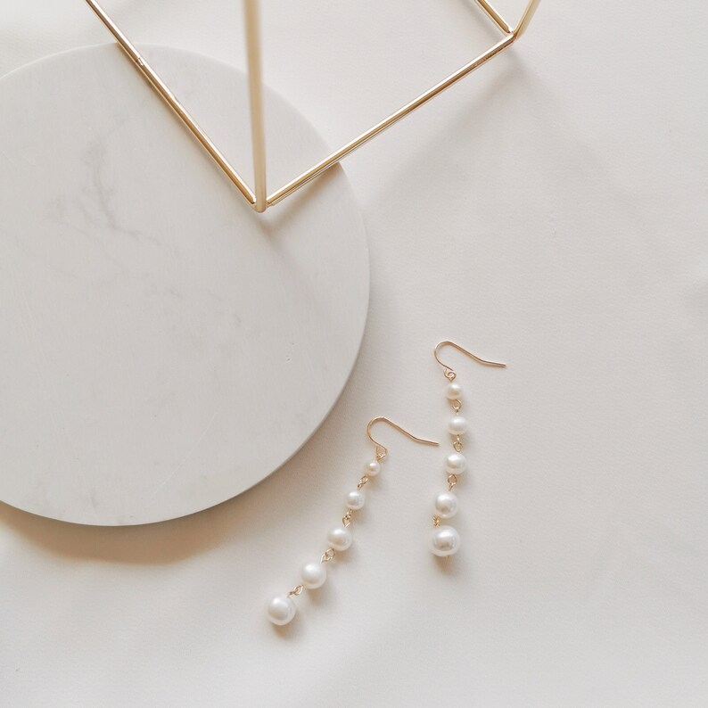 May include: A pair of gold dangle earrings with white pearl beads. The earrings are on a white surface.