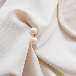 May include: Two white pearl earrings on a white fabric background.