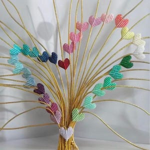 May include: A decorative arrangement featuring multiple heart-shaped ornaments in various colors, attached to golden-colored stems. The hearts are arranged in a bouquet shape, with a white heart at the base. The hearts are made of a textured material.