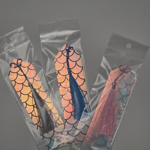May include: Three bookmarks in clear plastic sleeves, each featuring a holographic fish scale pattern in shades of orange, pink, and blue. Each bookmark has a hole at the top with a decorative thread tassel attached in grey, blue, or light purple.
