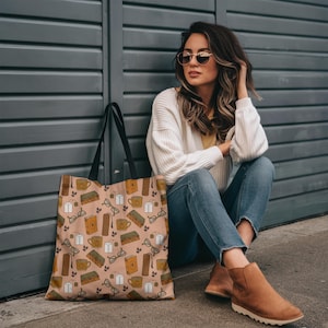 Könnte beinhalten: Beige Tragetasche mit einem sich wiederholenden Muster aus Büchern, Brillen, Kaffeetassen und Teebeuteln. Die Tasche hat schwarze Griffe und ist aus einem robusten Stoff gefertigt. Das Design ist in Braun-, Grün- und Weißtönen gehalten.
