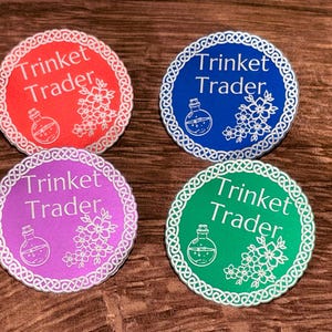 May include: Four circular coasters in red, blue, purple, and green, each featuring a white Celtic knot border, the text "Trinket Trader," a small potion bottle icon, and a cluster of floral blossoms printed in white on the colored background.