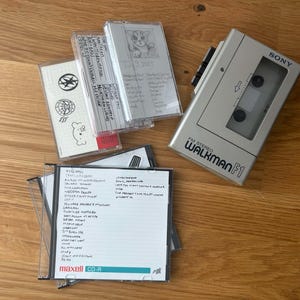 Puede incluir: Un reproductor de casetes estéreo FM Sony Walkman F1 plateado junto a varias cintas de casete y una funda de CD-R. Las cintas de casete tienen etiquetas y dibujos escritos a mano. La funda de CD-R tiene una lista de canciones escrita a mano.