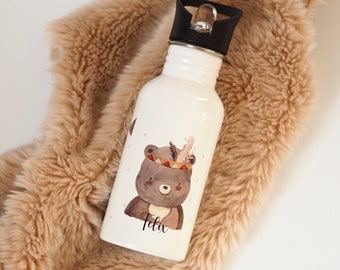 May include: A white water bottle with a black lid featuring a cartoon bear wearing a Native American headdress. The bear is brown with a watercolor style. The name "Felix" is written below the bear in black script. The bottle is resting on a tan, fluffy blanket.