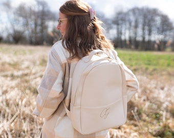 Könnte beinhalten: Ein cremefarbener Lederrucksack mit einem oberen Griff und einem goldenen Monogramm der Buchstaben "CG".