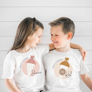 Peut inclure: Deux enfants portant des t-shirts blancs avec un graphique d'un cône scolaire et le texte "Endlich Schulkind" et leurs noms "Johanna" et "Karl".
