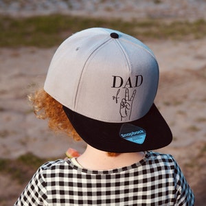 Könnte beinhalten: Ein Kind trägt eine graue und schwarze Baseballkappe mit dem Text "DAD of" und einem Peace-Zeichen. Die Kappe hat einen schwarzen Schirm und ein blaues "snapback"-Label. Das Kind trägt ein schwarz-weiß kariertes Hemd.