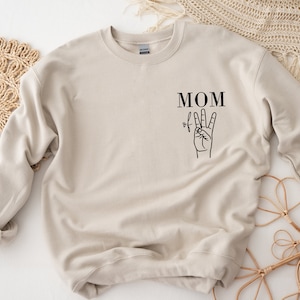 Könnte beinhalten: Ein beiger Pullover mit dem Text "MOM of" und einer Hand, die das Friedenszeichen macht.
