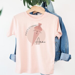 May include: Pink t-shirt with a graphic of a cartoon school cone with the text "Endlich Schulkind" and the name "Anni" printed on it.