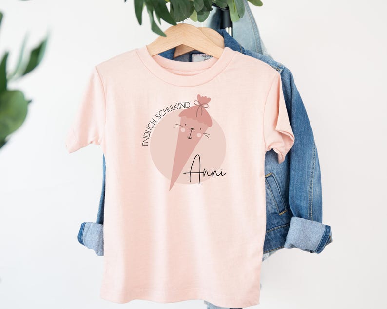 May include: Pink t-shirt with a graphic of a cartoon school cone with the text "Endlich Schulkind" and the name "Anni" printed on it.