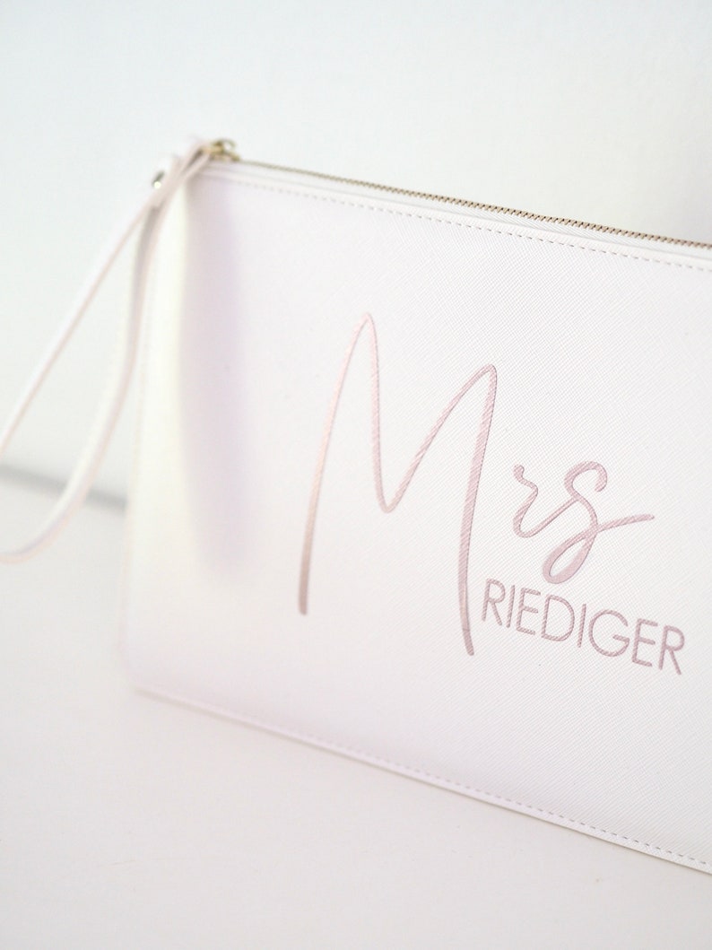 Pode incluir: Clutch branca com fecho de correr e uma al&ccedil;a de pulso. A bolsa tem a palavra "Mrs" e o sobrenome "RIEDIGER" bordado em rosa.