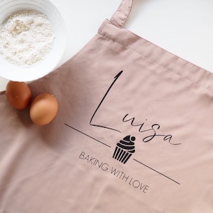 Könnte beinhalten: Eine rosa Schürze mit dem Text "Luisa Baking with Love" und einer Cupcake-Grafik.