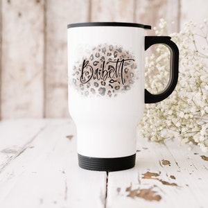 Op de afbeelding: Witte reistumbler met een zwarte deksel en handvat. De tumbler heeft een luipaardprint met de naam "Babett" in zwarte cursieve letters.