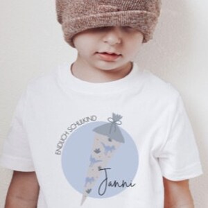 May include: White t-shirt with a blue circle and a drawing of a decorated cone with the text "Endlich Schulkind" and the name "Janni".