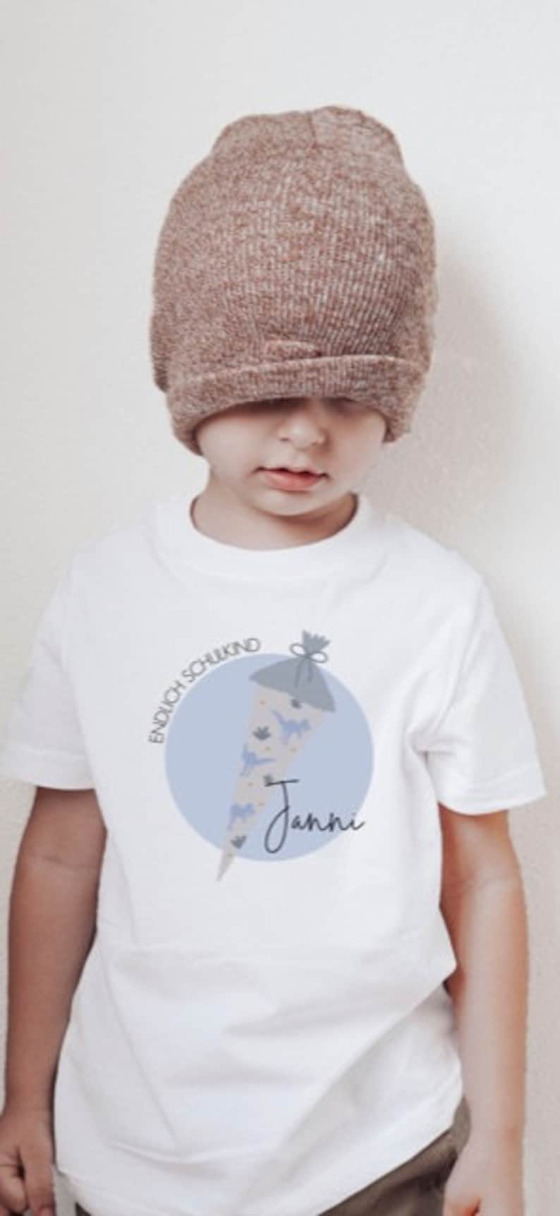 May include: White t-shirt with a blue circle and a drawing of a decorated cone with the text "Endlich Schulkind" and the name "Janni".