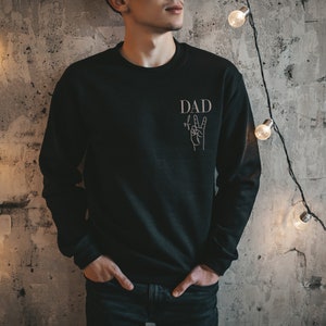 Könnte beinhalten: Ein schwarzer Pullover mit dem Wort "DAD" und einer Hand, die das Peace-Zeichen macht, auf der Vorderseite.