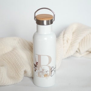 May include: White stainless steel water bottle with a bamboo lid and a carrying handle. The bottle has a floral design and the letter "B" in a beige color.