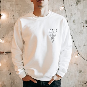 Könnte beinhalten: Weißer Pullover mit dem Text "DAD" und einer Hand, die das Friedenszeichen macht.