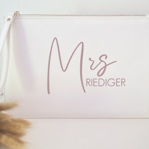 Pode incluir: Clutch branca com fecho de correr e uma al&ccedil;a de pulso. A bolsa tem o texto "Mrs. Riediger" impresso em rosa.