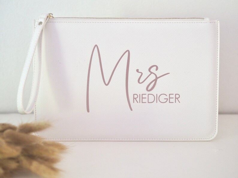 Pode incluir: Clutch branca com fecho de correr e uma al&ccedil;a de pulso. A bolsa tem o texto "Mrs. Riediger" impresso em rosa.