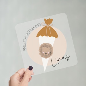 Könnte beinhalten: Eine durchsichtige Plastikkarte mit einer braunen und weißen Illustration eines Löwenkopfes in einem Kegel mit einer braunen Tüte oben drauf. Der Text "ENDLICH SCHULKIND" befindet sich auf der linken Seite der Karte und der Name "Linus" auf der rechten Seite.