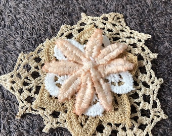 May include: A delicate crocheted flower with a peach-colored center and white lace trim. The flower is layered on top of a larger crocheted doily.