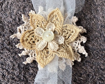 May include: A crocheted flower with a pearl button in the center, accented with a white tulle bow. The flower is beige and cream in color.