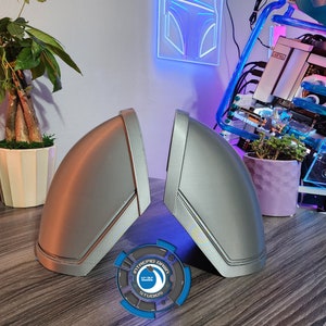 Pu&ograve; includere: Due coperture per altoparlanti stampate in 3D di colore argento con un design a strisce nere. Le coperture sono su un tavolo di legno con un logo blu e bianco al centro.