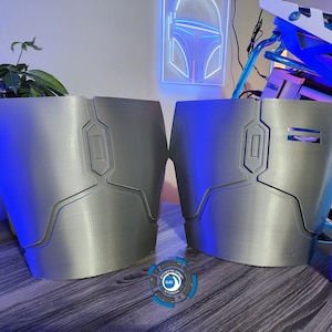 May include: Two silver-colored 3D printed pieces of armor with geometric designs. The pieces are on a wooden surface.