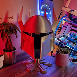 Puede incluir: Un casco impreso en 3D plateado y rojo con un soporte. El casco tiene una visera y una parte superior puntiaguda. El soporte es una base circular con un diseño que se asemeja a una estrella.