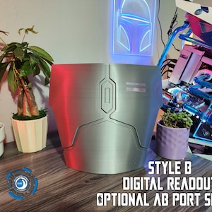 May include: A silver-colored 3D printed chest piece with a digital readout display and an optional port. The chest piece is on a wooden surface with a potted plant and a computer case in the background.