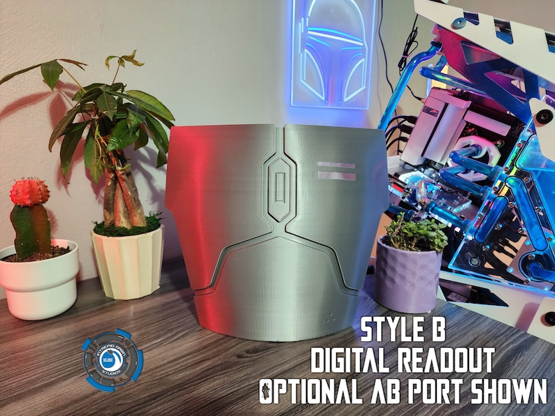 May include: A silver-colored 3D printed chest piece with a digital readout display and an optional port. The chest piece is on a wooden surface with a potted plant and a computer case in the background.