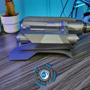 May include: A silver-colored, futuristic-looking prop weapon with a detailed design. The weapon is mounted on a black stand and has a logo on the stand that reads "Intrepid Design Studios".