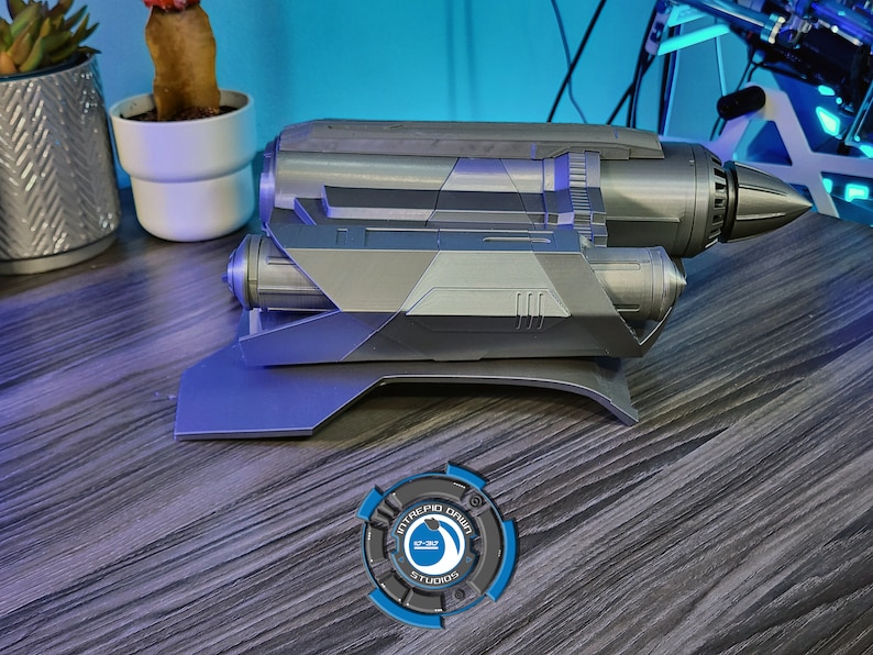 May include: A silver-colored, futuristic-looking prop weapon with a detailed design. The weapon is mounted on a black stand and has a logo on the stand that reads "Intrepid Design Studios".