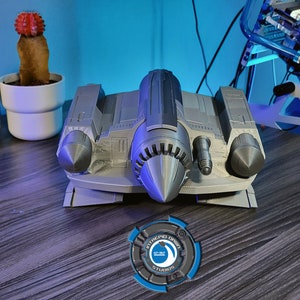 May include: A gray 3D printed model of a spaceship with a pointed nose and two side engines. The model is sitting on a wooden surface with a blue and gray logo in the foreground.