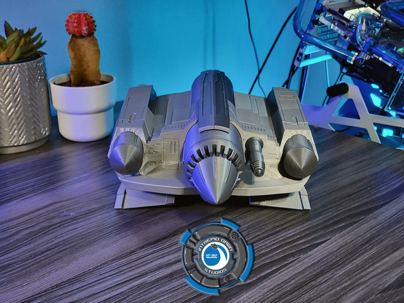 May include: A gray 3D printed model of a spaceship with a pointed nose and two side engines. The model is sitting on a wooden surface with a blue and gray logo in the foreground.