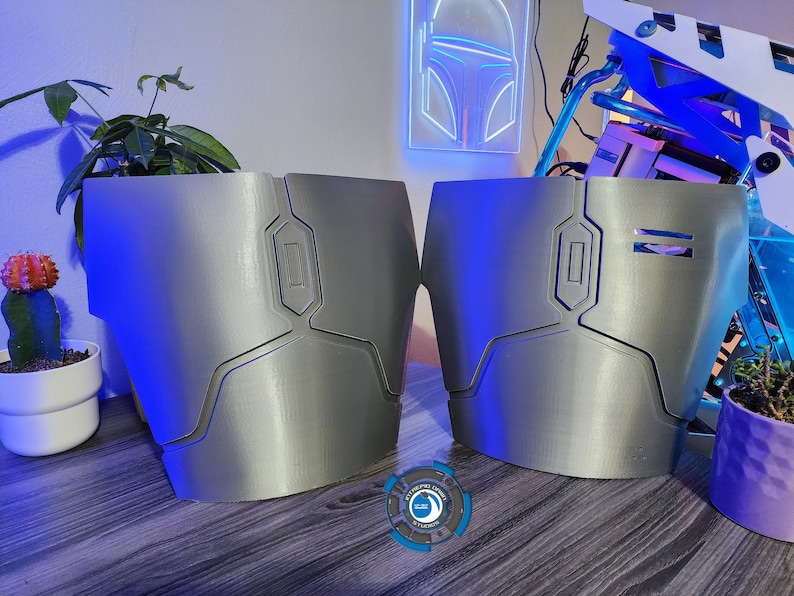 May include: Two silver-colored 3D printed pieces of armor with geometric designs. The pieces are on a wooden surface.