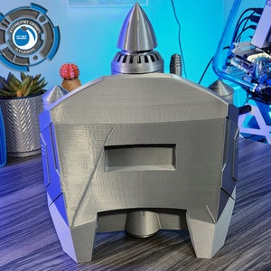 May include: A gray 3D printed model of a futuristic spaceship with a pointed nose and a rectangular window. The model is sitting on a wooden surface.