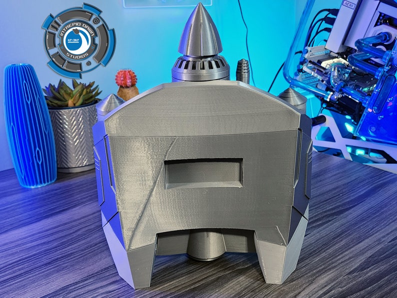 May include: A gray 3D printed model of a futuristic spaceship with a pointed nose and a rectangular window. The model is sitting on a wooden surface.