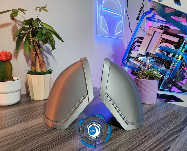 Pu&ograve; includere: Due coperture per altoparlanti stampate in 3D di colore argento con un design a strisce nere. Le coperture sono su un tavolo di legno con un logo blu e bianco al centro.