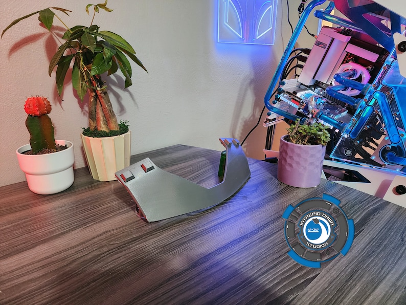 Op de afbeelding: Een grijs 3D-geprint object met een afgeronde vorm en twee kleine rechthoekige knoppen aan de bovenkant. Het object staat op een houten tafel met een roze cactus in een witte pot, een groene plant in een beige pot en een paarse plant in een witte pot. De achtergrond bevat een blauwe en witte computerbehuizing met een blauwe lichtstrip.
