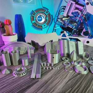 May include: A set of 3D printed model pieces in a silver color. The pieces are various shapes and sizes, including cylinders, cones, and rings. The pieces are arranged on a wooden surface.