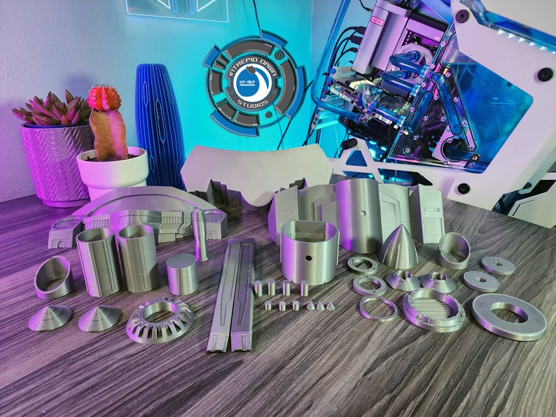 May include: A set of 3D printed model pieces in a silver color. The pieces are various shapes and sizes, including cylinders, cones, and rings. The pieces are arranged on a wooden surface.