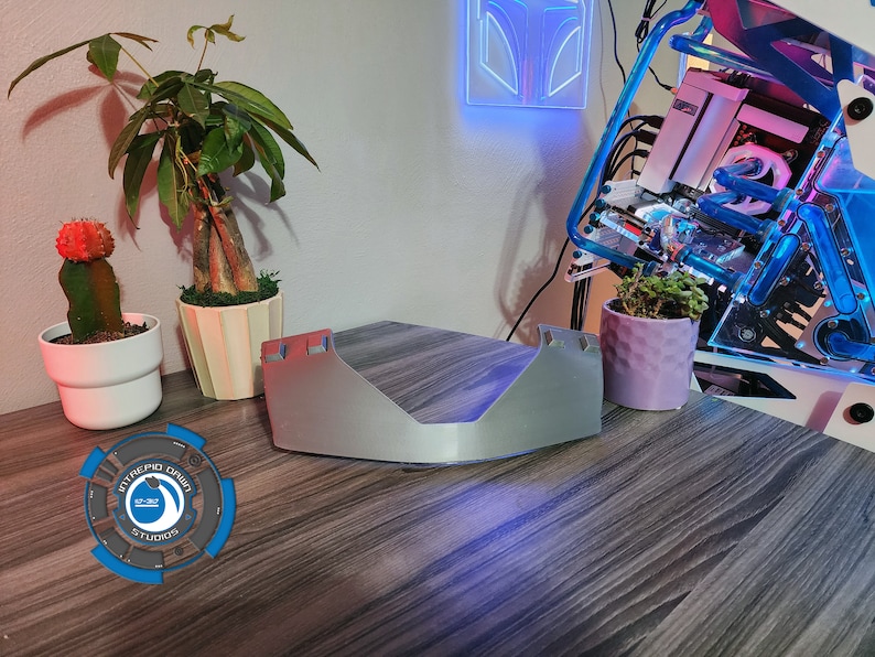 Op de afbeelding: Een zilverkleurig computeronderdeel met een gebogen ontwerp ligt op een houten tafel. Op de achtergrond staan twee planten in potten en een computerbehuizing met blauwe en paarse lichten.