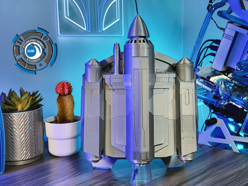 May include: A 3D printed model of a spaceship with a gray metallic finish. The spaceship has a pointed top and multiple engines on the bottom. The model is sitting on a clear glass base.