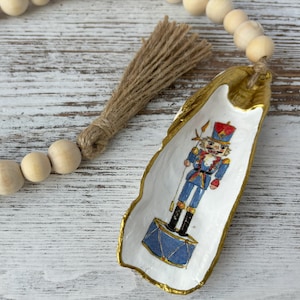 May include: A gold-painted oyster shell ornament with a nutcracker illustration. The nutcracker is wearing a blue and red uniform and holding a drum.