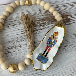 May include: A white, seashell-shaped ornament with a gold trim, featuring a hand-painted illustration of a nutcracker soldier in a blue and red uniform. The ornament is attached to a wooden bead garland with gold accents.